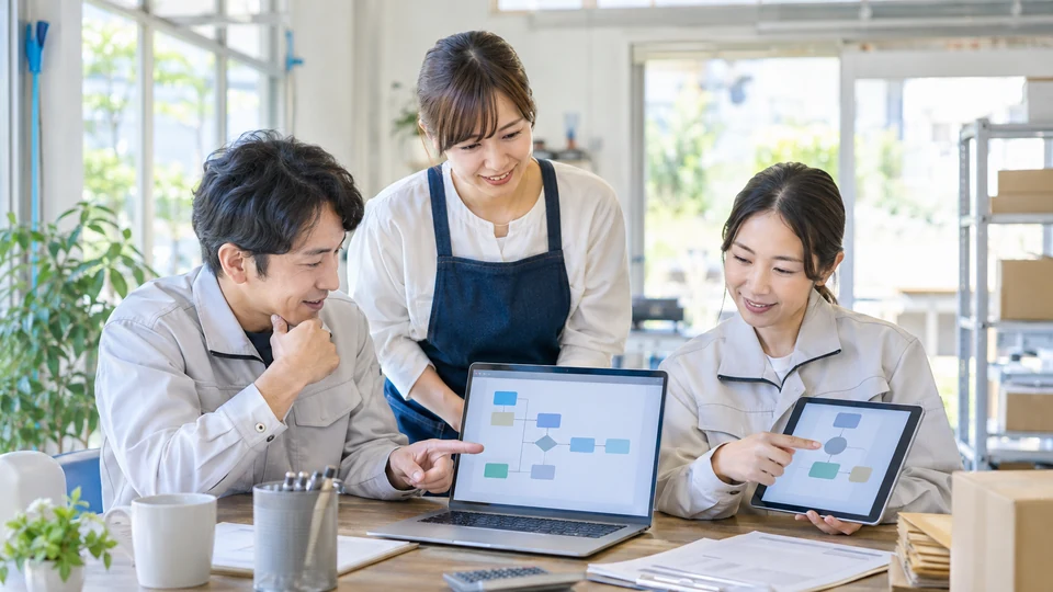 小規模事業の現場担当者が、ノートPCとタブレットで無文字の業務フロー画面を確認し、紙の作業をデジタル化する場面。AIやデジタル活用の検討を主題にする。